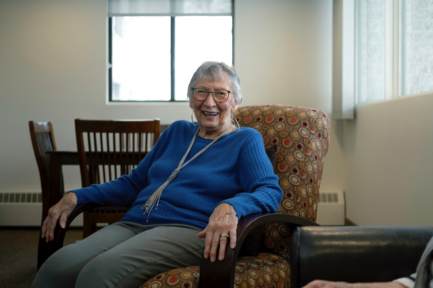 Smiling senior woman in a blue sweater sits in a patterned armchair in a light-filled room.