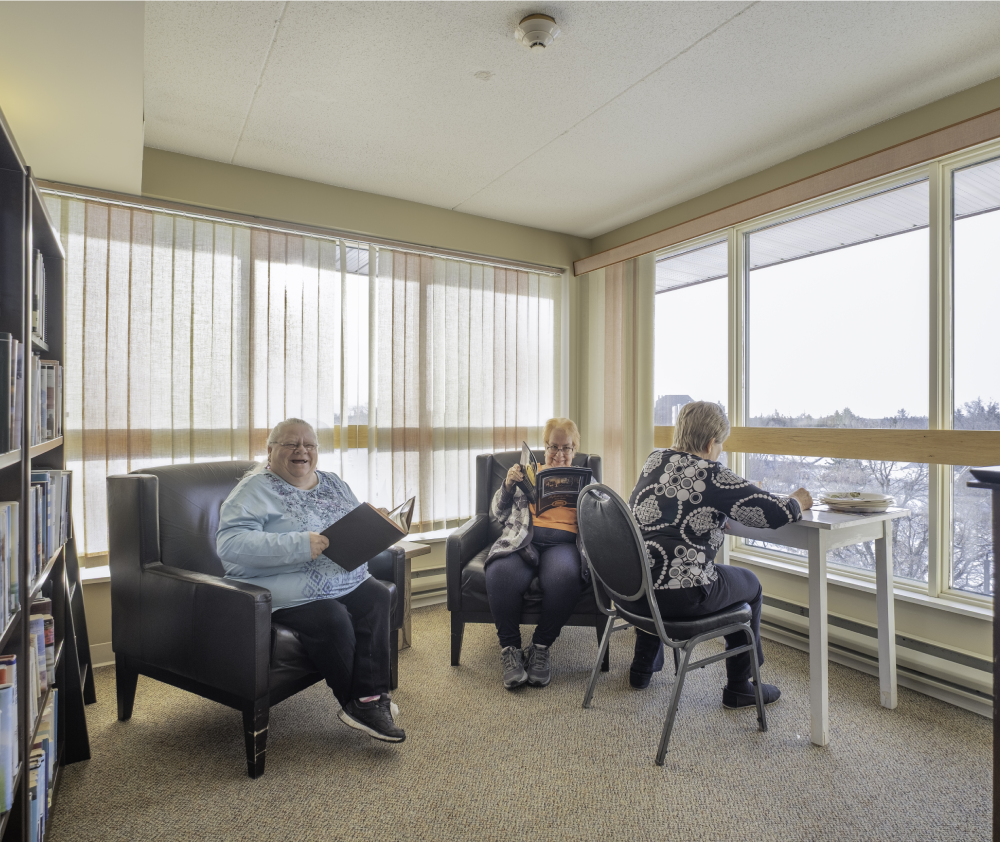 Three senior women relax in a community room, reading books and magazines, with a view of the outdoors. Three senior women relax in a community room, reading books and magazines, with a view of the outdoors.